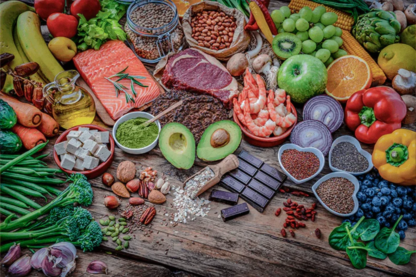 Overhead spread of whole foods including salmon, beef steak, shrimp, avocado, vegetables, fruit, nuts, and seeds on a wooden table